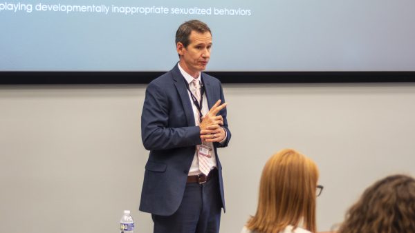 man in suit, talking to group, standing in front of presentation on screen